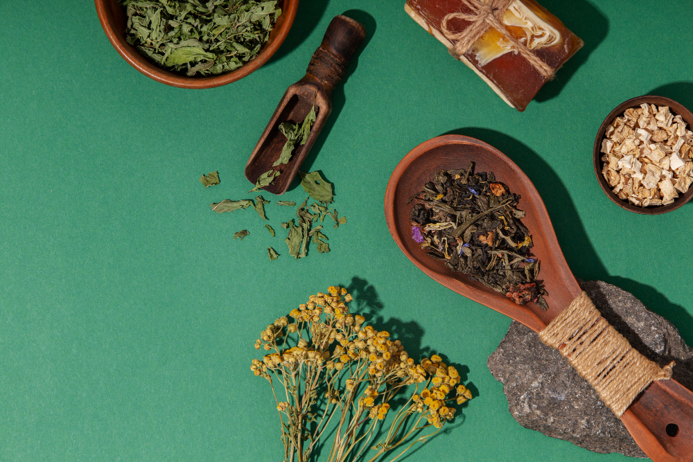 2 colheres de madeira cheia de ervas, 2 potes cheios de ervas de madeira com ervas em cima de uma mesa verde. Ao lado um ramo de flores amarelas